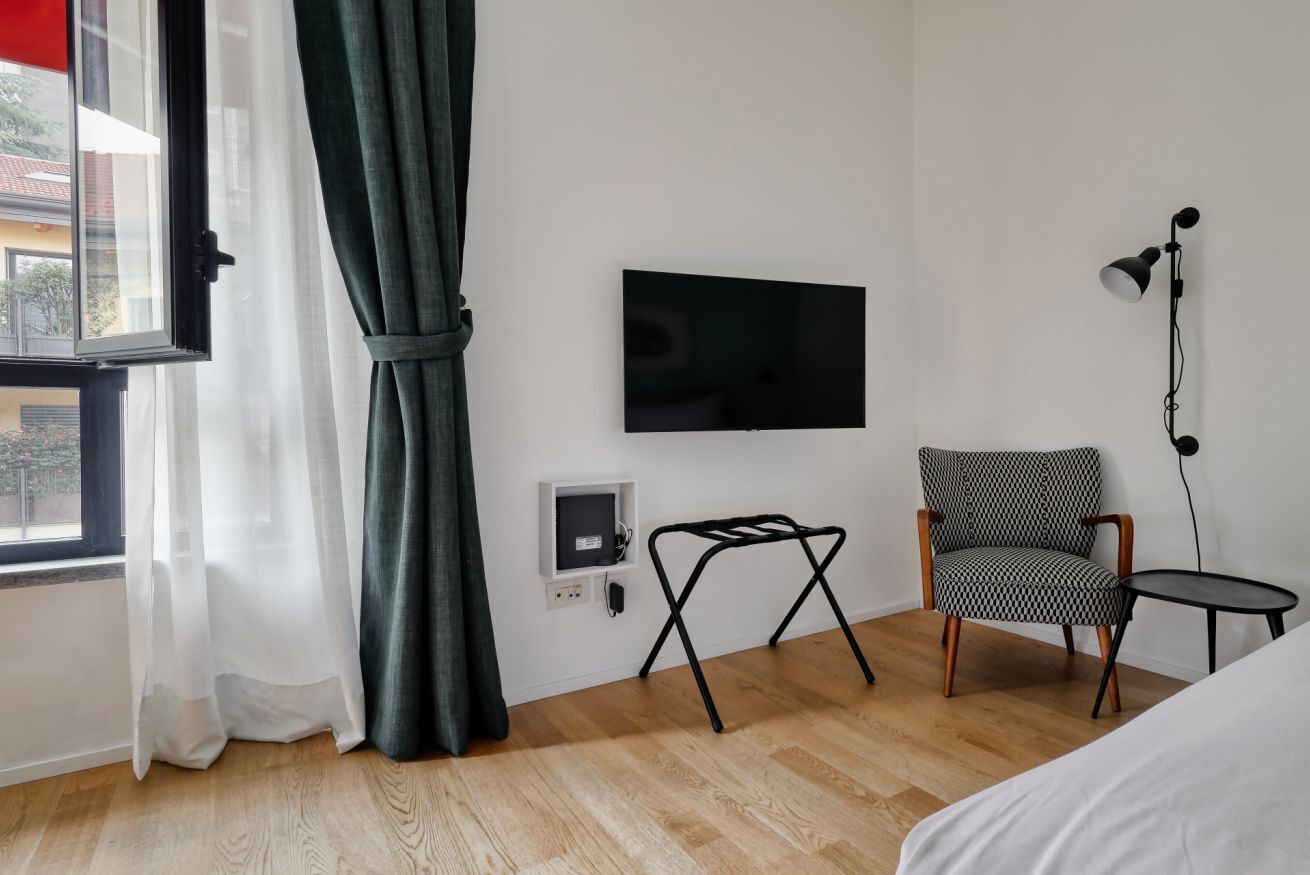 A hotel room with a bed, a TV mounted on the wall, a chair, a small table, and a window with curtains.