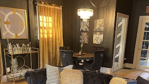 Un salón con un sofá de color oscuro, una mesa de centro redonda y una araña. Hay una mesa de comedor con sillas y una pared con paneles decorativos.