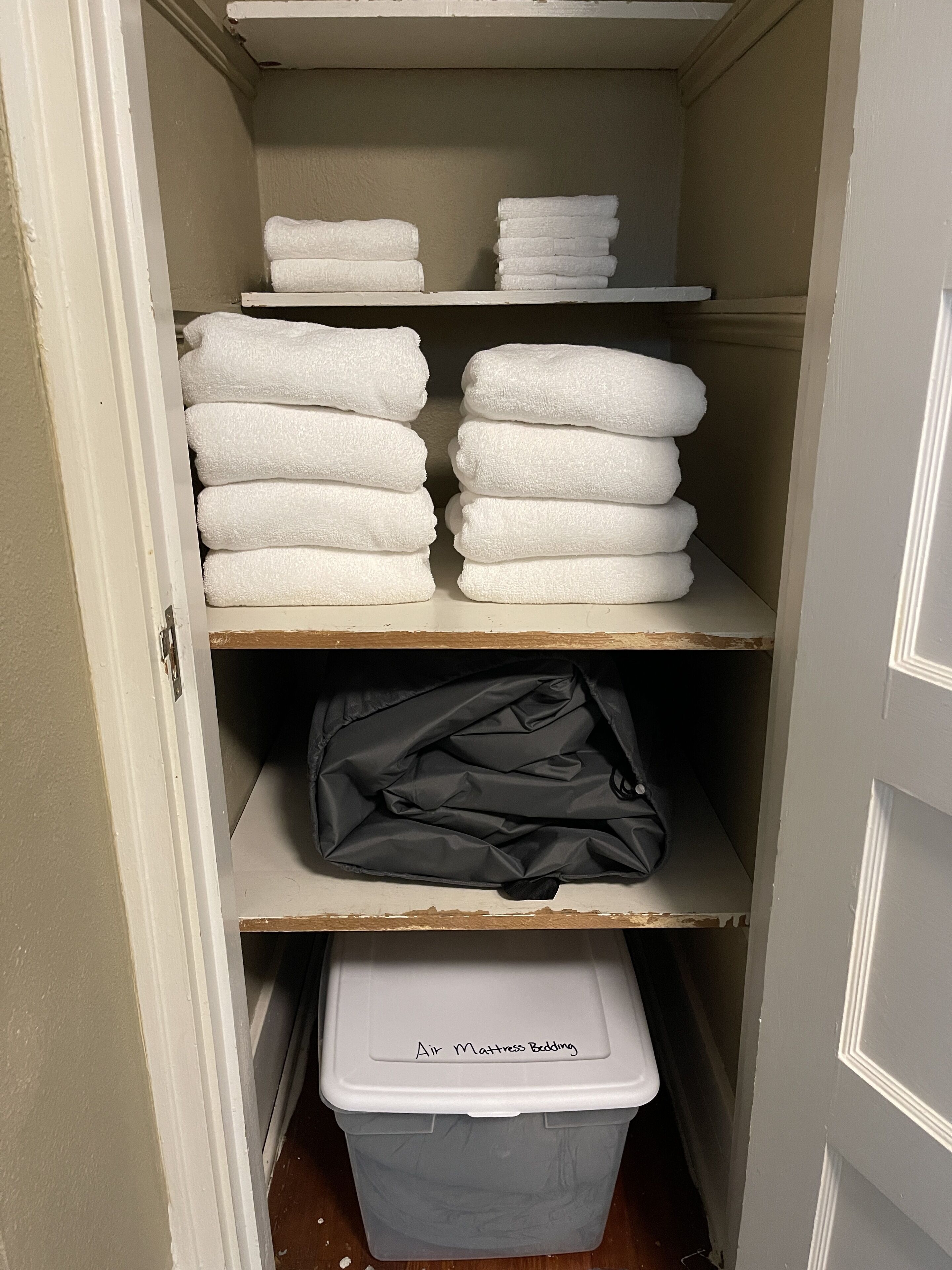 Un placard de rangement avec des étagères où sont pliés des serviettes blanches et un récipient en plastique gris étiqueté « Linge de lit pour matelas gonflable ».