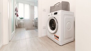 A modern laundry room with a front-loading washing machine, a small table with chairs, and a glass door leading to a bathroom. - Numero 11 Piazza Amendola - My Home in Como (Como)