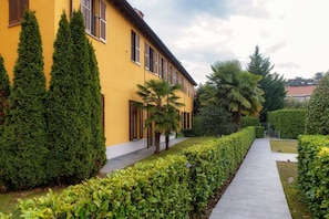 A yellow building with a paved walkway, neatly trimmed hedges, and tall evergreen trees. - Medel - My Home in Como (Appiano Gentile)
