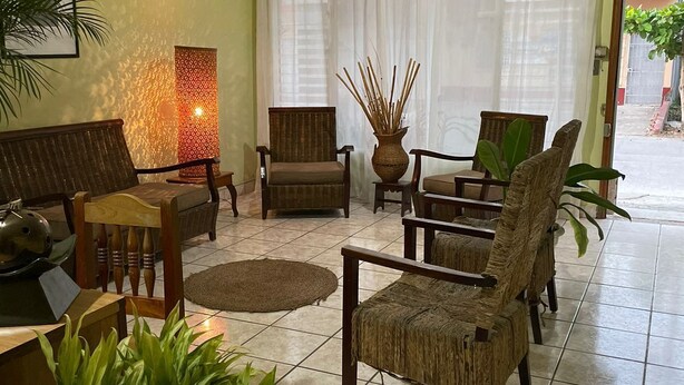 A living room with a ceiling fan, a large window with sheer curtains, and several wicker chairs. - Nakum Hotel (Flores)