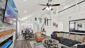A spacious living room with a fireplace, a large TV mounted on the wall, a ceiling fan, and a dining area in the background.