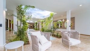 A spacious indoor area with a high ceiling, featuring a central pathway, white wicker chairs, a small table, and various indoor plants. - Casa Mangos Casa de Campo DR  (La Romana)