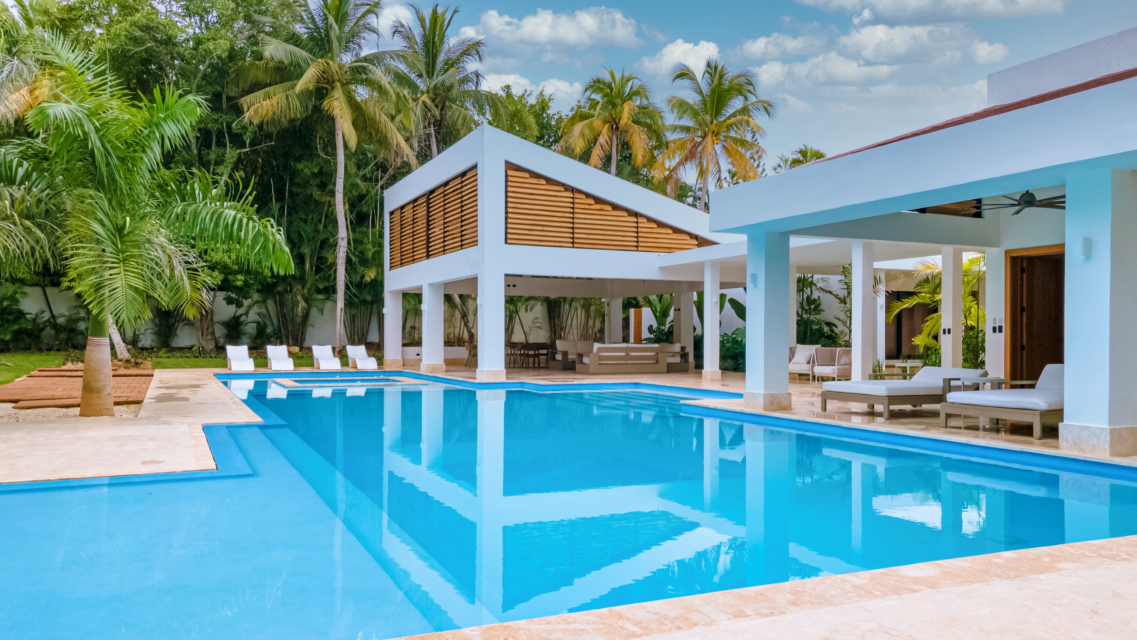A modern pool area with a clear blue pool, lounge chairs, and a covered seating area surrounded by palm trees.