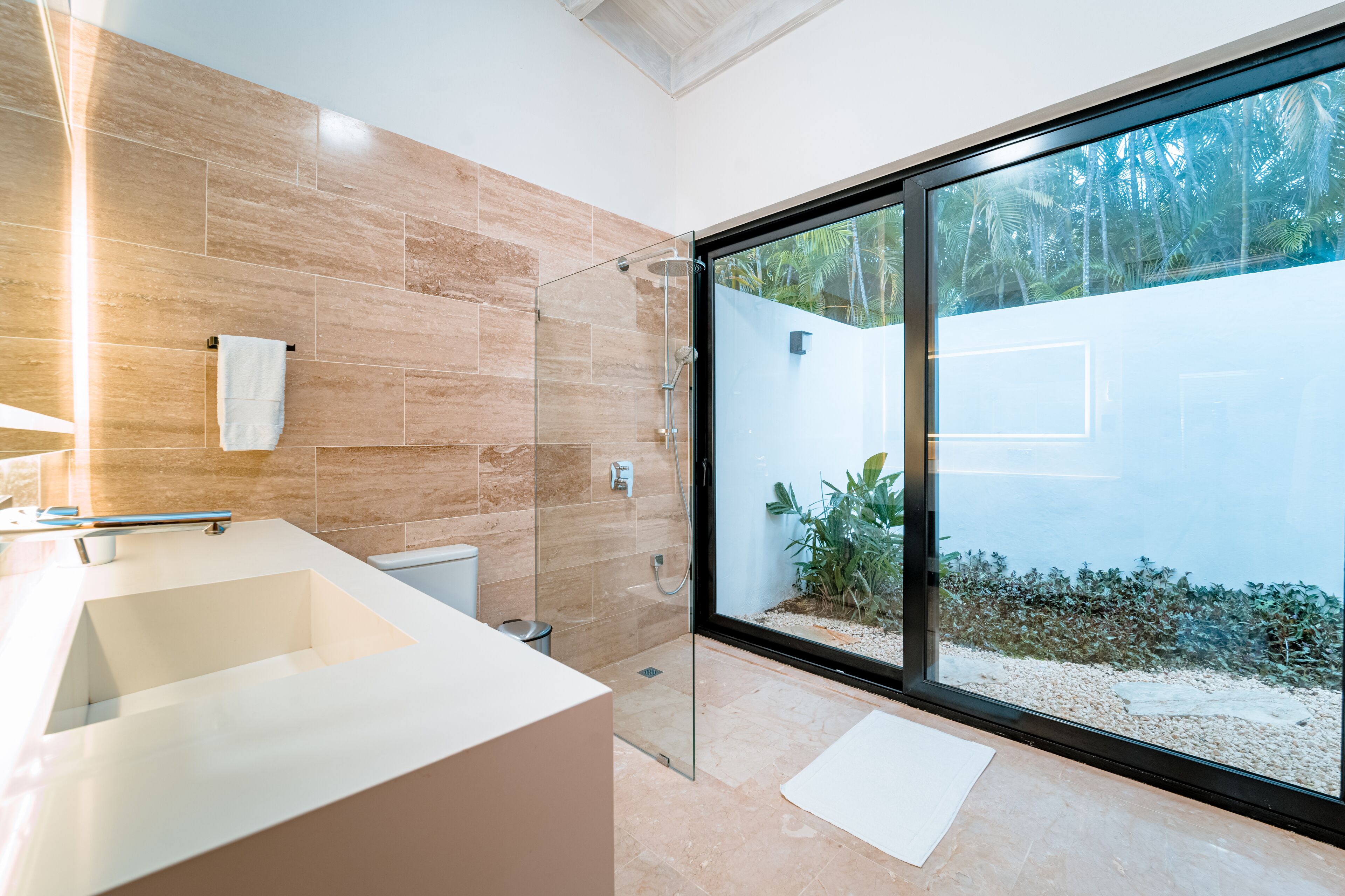 Un baño moderno con una ducha de vidrio amplia, un lavabo blanco y vistas a un jardín tropical a través de la puerta de cristal.