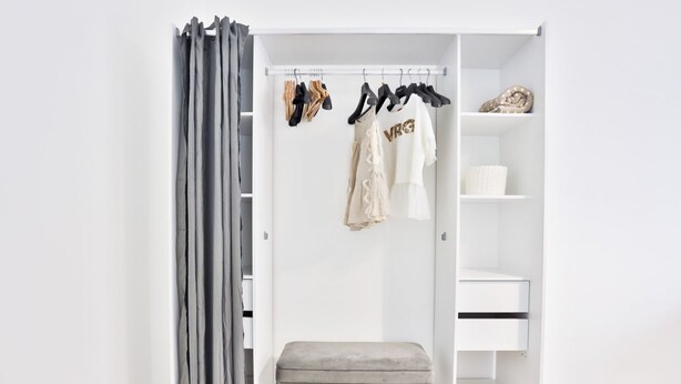 A walk-in closet with a grey bench, hanging clothes, and shelves.