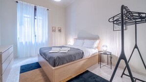Un dormitorio con una cama, una mesita de noche con lámpara, una ventana con cortinas y un tendedero en la pared.