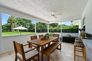 A covered patio with a wooden table and chairs, a ceiling fan, and large windows offering a view of a garden.