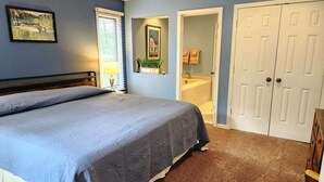 Un dormitorio con cama, mesita de noche, lámpara, ventana con persianas, baño con lavamanos y espejo, y armario.