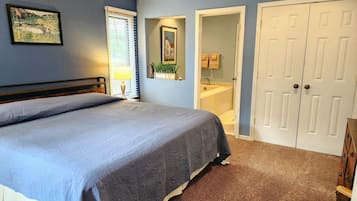 A bedroom with a bed, a nightstand, a lamp, a window with blinds, a bathroom with a sink and mirror, and a wardrobe.