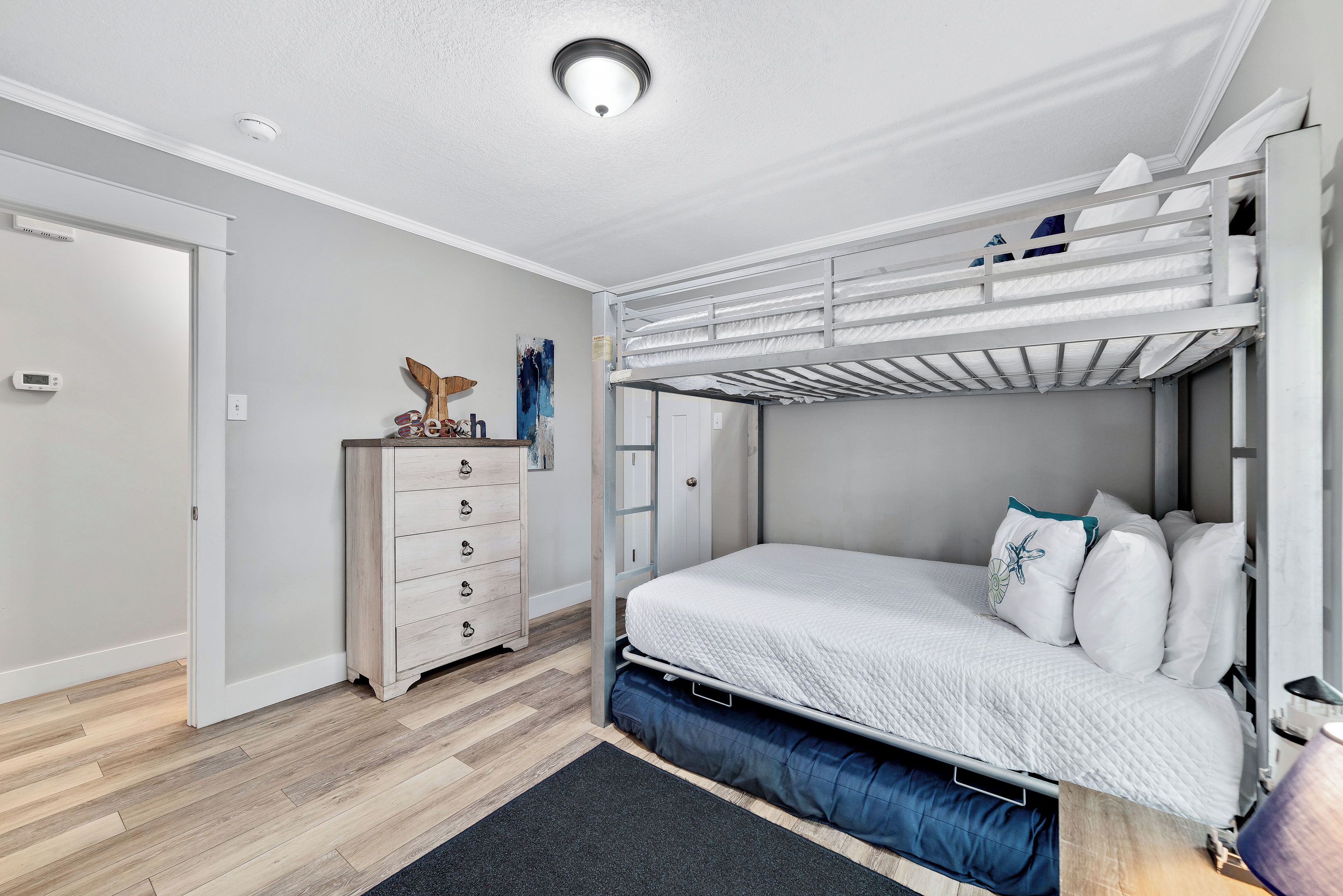 Un lit superposé avec un matelas et des oreillers blancs, une commode avec des tiroirs, une petite table avec une lampe et un luminaire au plafond.