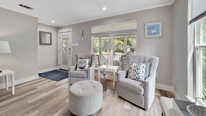 Un amplio salón con un sofá gris, un reposapiés y un sillón, una mesita blanca y un gran ventanal con persianas.
