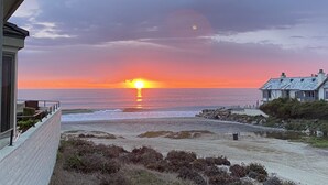 Une vue sur le coucher de soleil sur la côte, prise depuis un balcon donnant sur une plage, avec une maison et un promontoire rocheux.