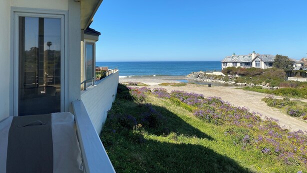 Une propriété située sur la côte, dotée d’un balcon donnant sur la mer, d’un jardin agrémenté de fleurs violettes et offrant une vue sur d’autres maisons au loin.