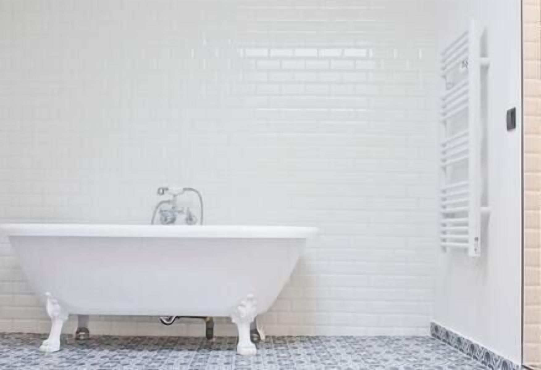 Een wit vrijstaand bad in een badkamer met witte betegelde muren en een vloer met een patroon.