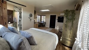 Quarto com uma cama grande, uma escrivaninha e uma cadeira. Há uma janela com persianas, uma porta e uma planta.