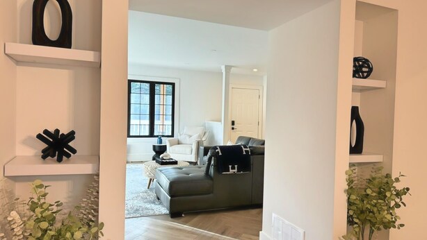 Una sala moderna con un sofá gris, un sillón blanco y una mesa de centro negra. Hay jarrones negros decorativos en estanterías blancas y una planta en maceta al frente.