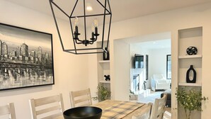 Un comedor con un tazón negro sobre una mesa, sillas de madera, una lámpara con diseño geométrico y un cuadro de un paisaje urbano en la pared.