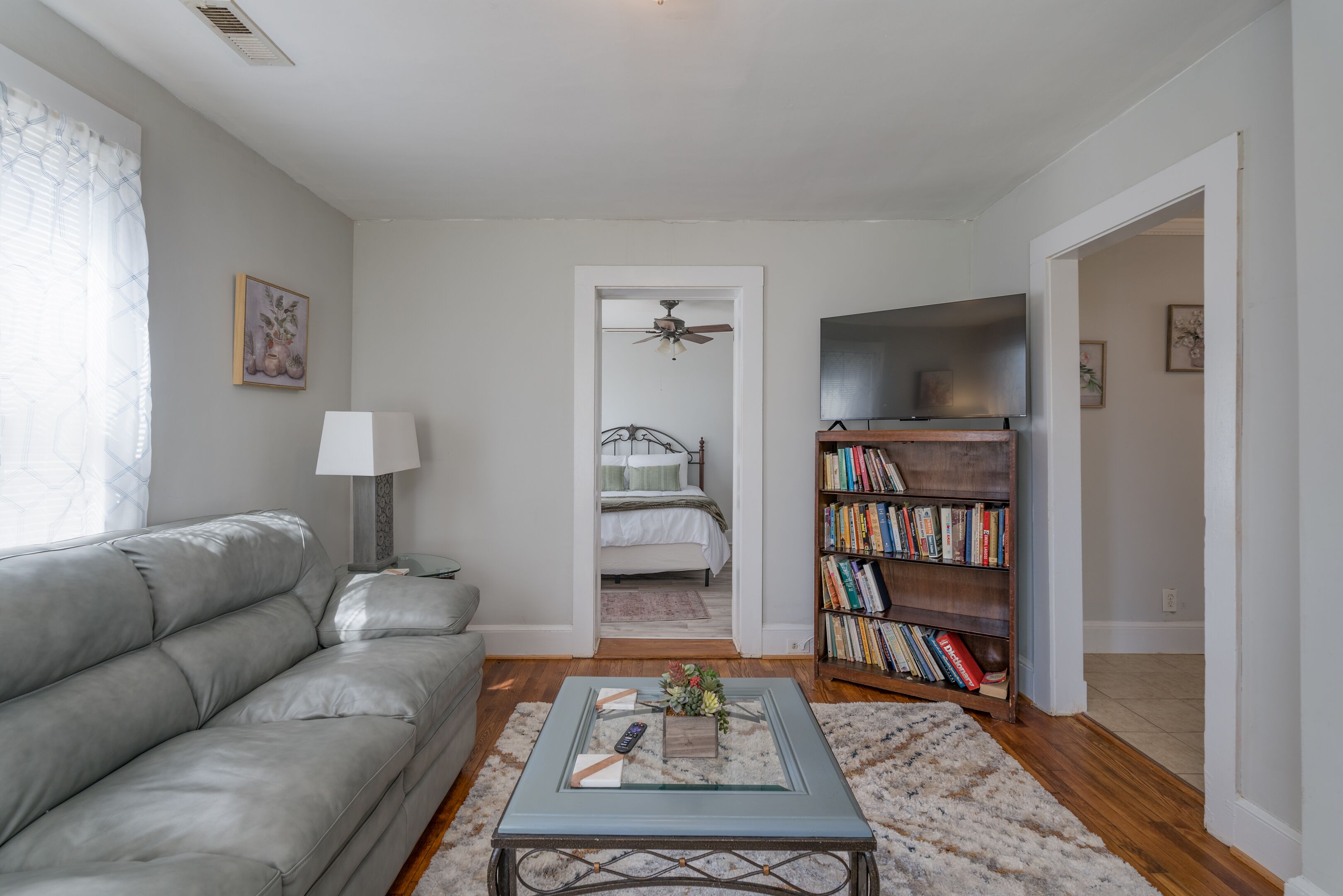 A living room with a grey sofa, a glass coffee table, a bookshelf, and a door leading to a bedroom.