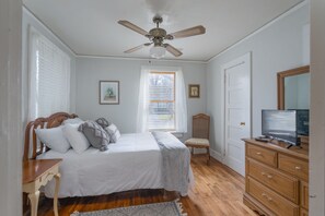 A bedroom with a bed, a wooden dresser, a television, a ceiling fan, and a window with blinds.