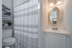 A bathroom with a striped shower curtain, a toilet, a shelf with folded towels, a sink with a mirror, and a light fixture above the mirror.