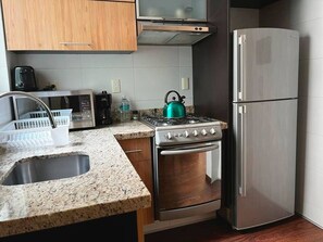 A kitchen with a granite countertop, stainless steel appliances, a microwave, a kettle, and a refrigerator.