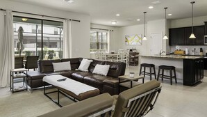 A modern living room with a brown leather sofa, a dining area with a kitchen island, and large windows.