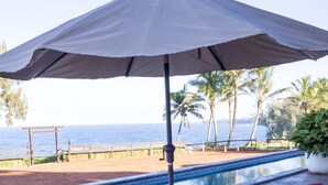 A poolside area with two wooden lounge chairs under a large umbrella, a pool, and a view of the ocean and palm trees.