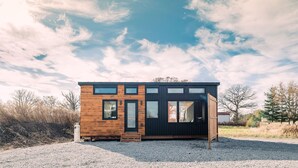 Une cabane moderne, dotée d’une façade en bois et de grandes fenêtres, située dans un cadre naturel, avec un sol de gravier et des arbres en arrière-plan.