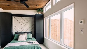 Une chambre avec une grande fenêtre, un lit avec une literie verte, un ventilateur de plafond et un plafond en bois.