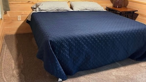A bedroom with a wooden ceiling, a bed with a blue bedspread, two patterned pillows, a nightstand with a lamp, and a carpeted floor. - Cabin 2 at Timberlane Resort on Long Lake (Park Rapids, MN)