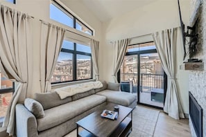 Una sala de estar con sofá, mesa de centro y ventanas grandes con cortinas.