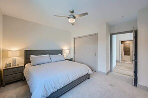 Un dormitorio con cama, mesitas de noche, ventilador de techo y una puerta que da a otra habitación.