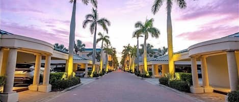 A pathway lined with palm trees and covered structures, leading towards a building.