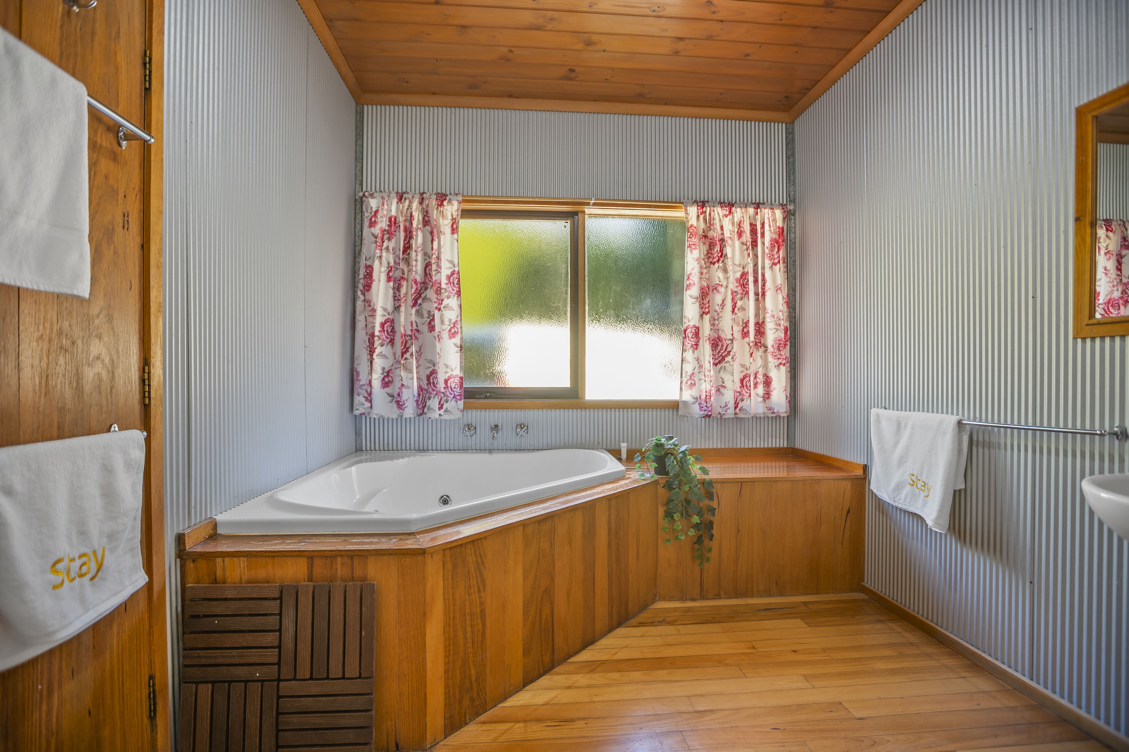 A bathroom with a wooden bathtub, a window with floral curtains, and a towel rack with a towel saying "Stay".