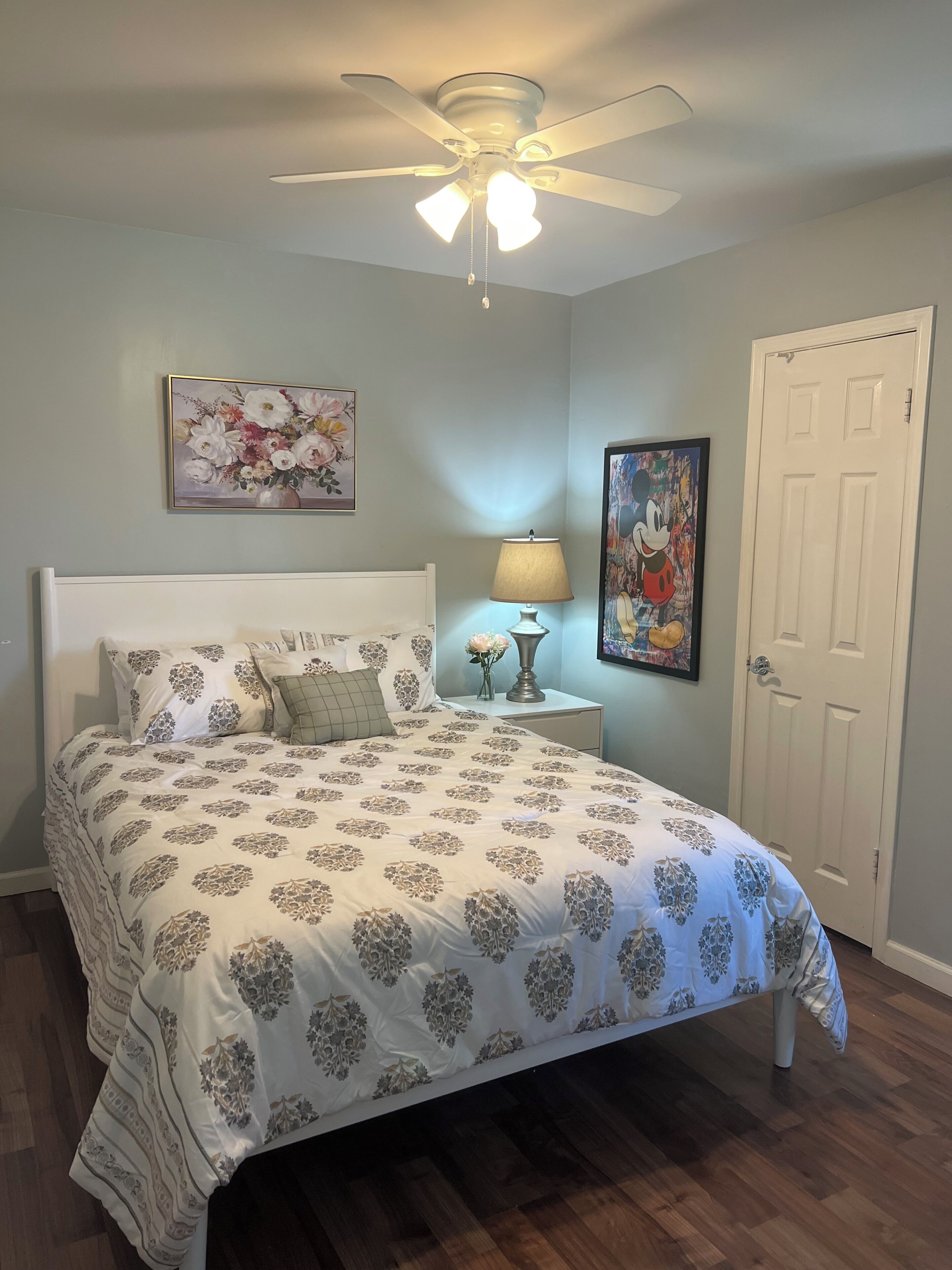 A bedroom with a bed, a nightstand, a lamp, a ceiling fan, a door, and two framed pictures on the wall.