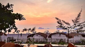 A seaside restaurant with wooden tables and chairs, a thatched roof, and a view of the ocean and distant islands.