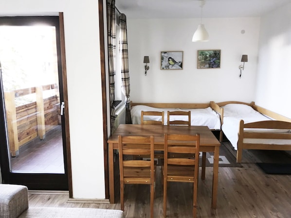 A cozy room with a wooden table and chairs, a sofa, and a bed. There are two wall-mounted lamps and two framed pictures.