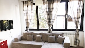 A living room with a sectional sofa, a flat-screen TV, a red cabinet, and large windows with plaid curtains.