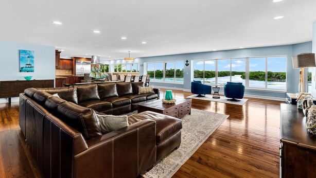 A spacious living room with a large brown leather sofa, a blue armchair, and a wooden coffee table. The room has large windows offering a view of the ocean.