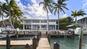 A wooden dock leading to a white building with a balcony, surrounded by palm trees and a clear sky.