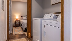 Un cuarto de lavandería con lavadora y secadora, una cama con mesita de noche y lámpara, y una cita enmarcada en la pared.