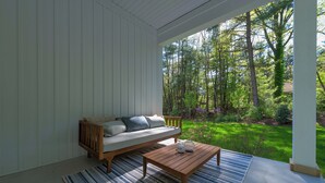 Un porche cubierto con un banco de madera y una mesa de centro, un tapete azul y blanco a rayas, y vistas a un exuberante jardín verde.