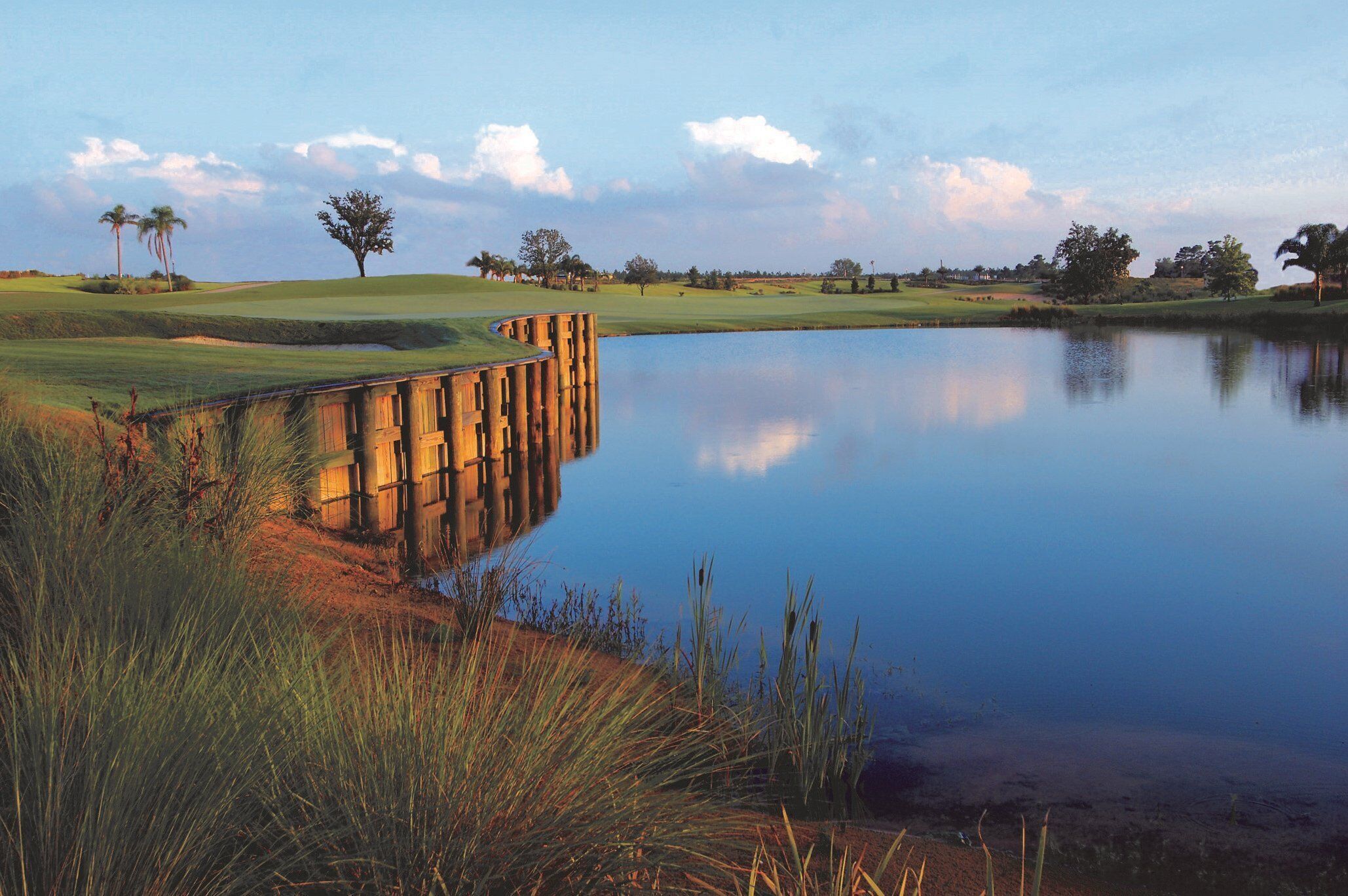 A tranquil golf course with a wooden bridge over a calm pond, surrounded by grass and trees.
