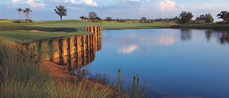 A tranquil golf course with a wooden bridge over a calm pond, surrounded by grass and trees.