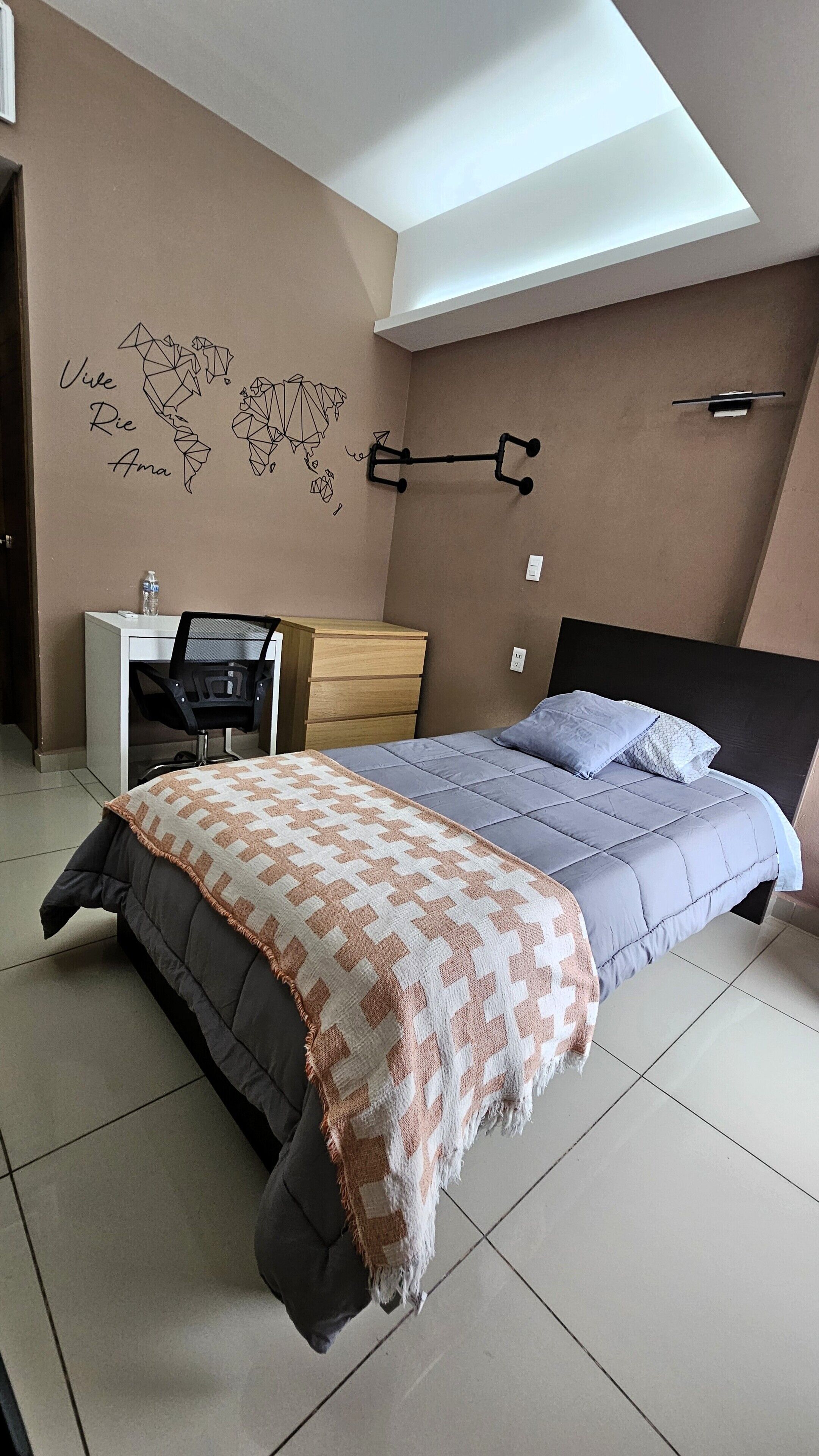 Un dormitorio con cama, escritorio y silla. Hay un mapa mural y una lámpara.