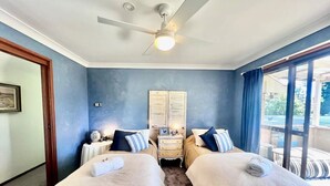 Una habitación con dos camas, un ventilador de techo, una mesita de noche y un ventanal grande con cortinas.