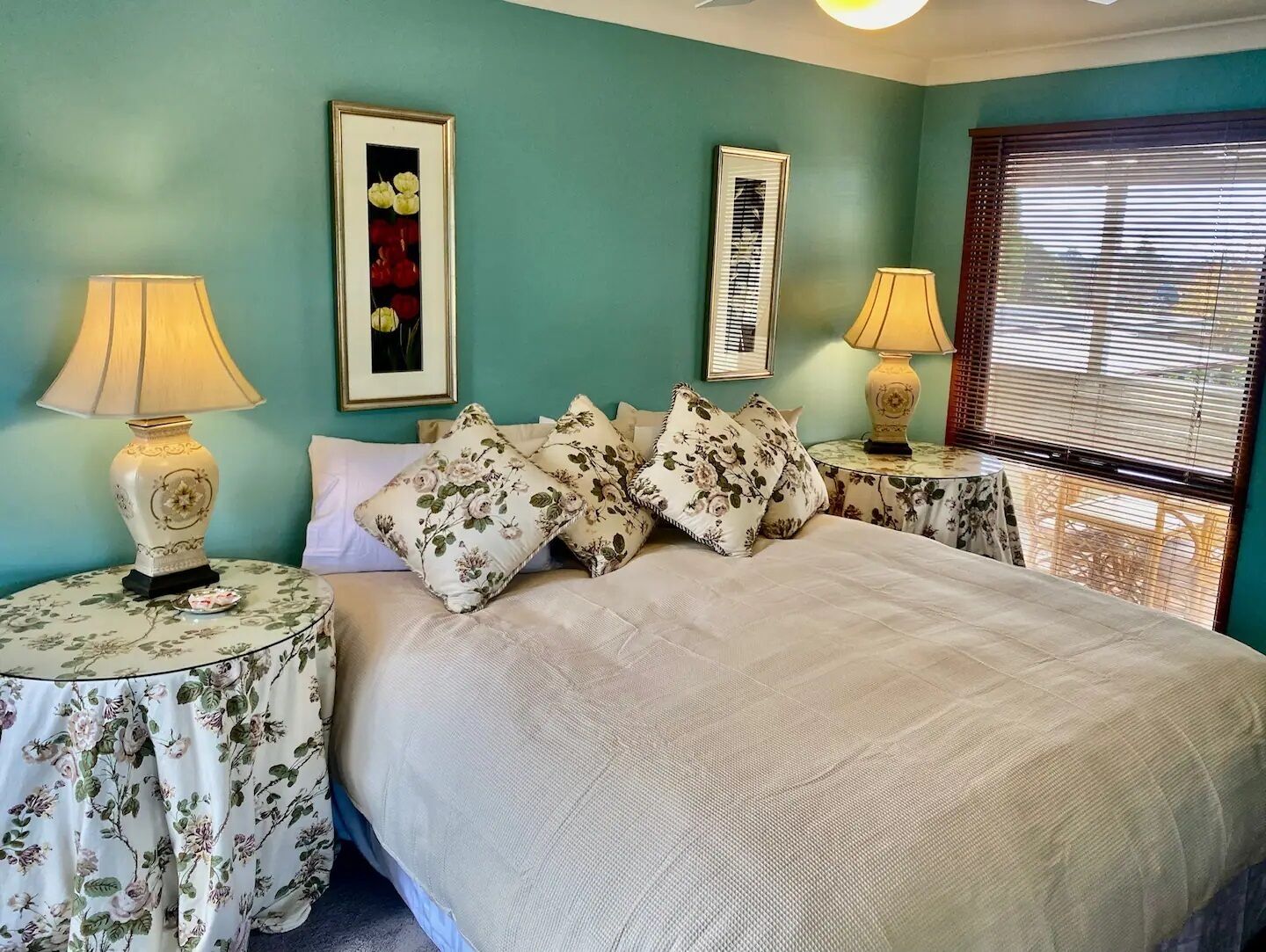 Une chambre à coucher avec un grand lit, des coussins à motifs floraux, deux tables de chevet avec des lampes et une fenêtre avec des stores.