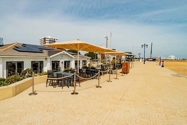 A seaside restaurant with outdoor seating, a white building, and a sandy beach in the background.